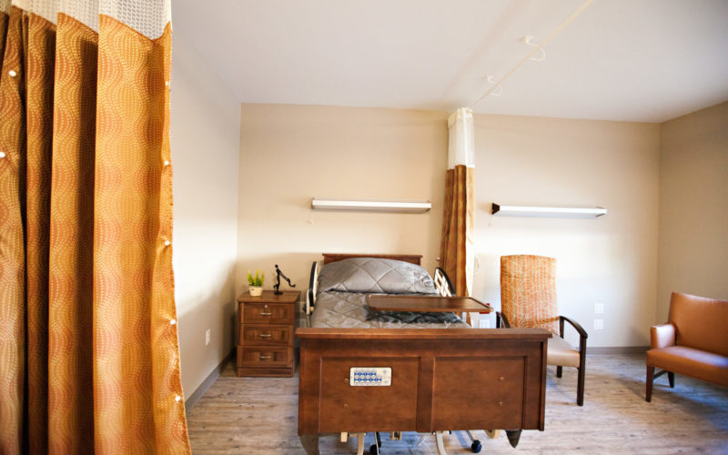 A resident room with one bed, a tray, a nightstand and two soft chairs. A large curtain hangs from the ceiling and can be pulled for privacy.
