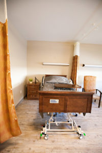 A resident room with a bed, tray, nightstand and a soft chair. A large curtain hangs from the ceiling and can be pulled closed for privacy.