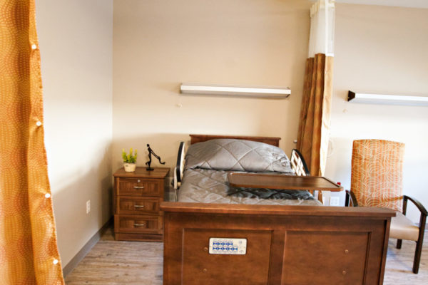 A resident room with a bed, tray, nightstand and a soft chair. A large curtain hangs from the ceiling and can be pulled closed for privacy.