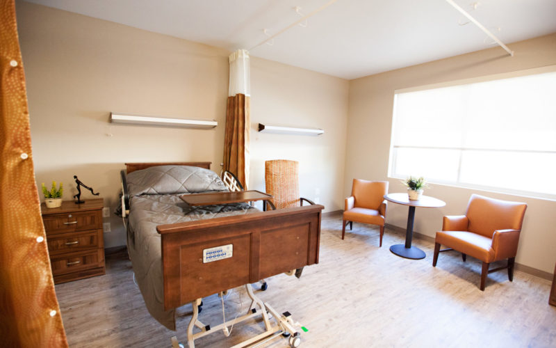 A resident room with a bed, tray, three soft chairs and a small round table sitting in front of a large window. A large curtain hangs from the ceiling and can be closed for privacy.
