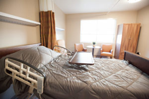 A resident room with a bed, tray, three soft chairs and a small round table sitting in front of a large window a clothes cabinet is next to the chairs. A large curtain hangs from the ceiling and can be pulled closed for privacy.