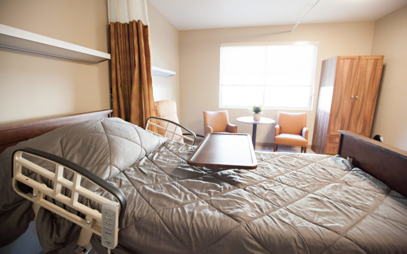 A resident room with a bed, tray, three soft chairs and a small round table sitting in front of a large window a clothes cabinet is next to the chairs. A large curtain hangs from the ceiling and can be pulled closed for privacy.