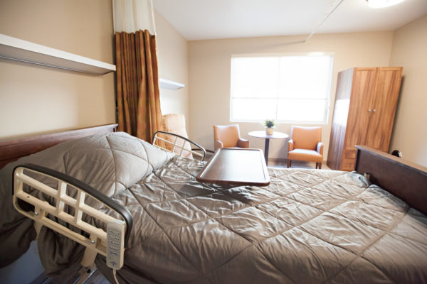 A resident room with a bed, tray, three soft chairs and a small round table sitting in front of a large window a clothes cabinet is next to the chairs. A large curtain hangs from the ceiling and can be pulled closed for privacy.