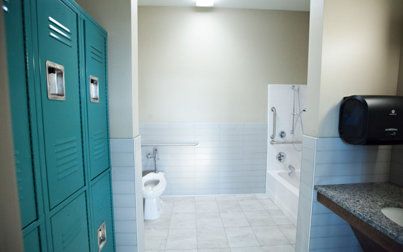 A tile floored bathroom with two rows of lockers on the left and a sink with a counter across from them. A toilet and tub with grab bars are at the far end of the room.