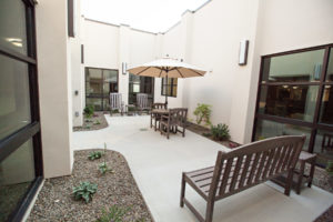 A concrete sidewalk with rocking chairs, a table with an umbrella and chairs, and a bench are placed along the sidewalk. Flower beds are on either side of the sidewalk. The area is between two walls and windows of a stucco building.