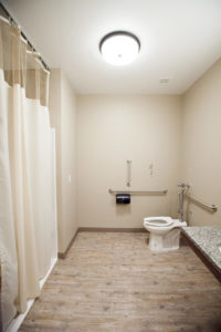A narrow bathroom with a counter, toilet and shower. Grab bars are placed on the wall throughout the room.