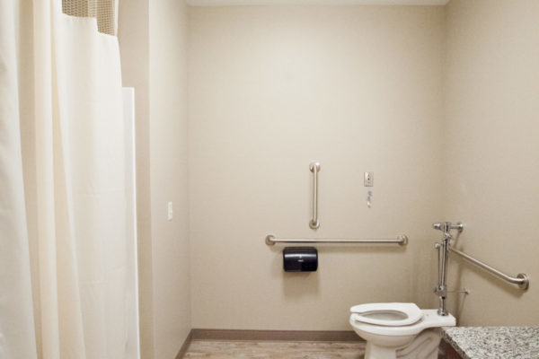 A narrow bathroom with a counter, toilet and shower. Grab bars are placed on the wall throughout the room.