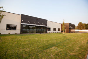 A large grass area with two deciduous trees that leads to a rock bed with shrubs then to a one-story, flat-roofed building with lots of windows.