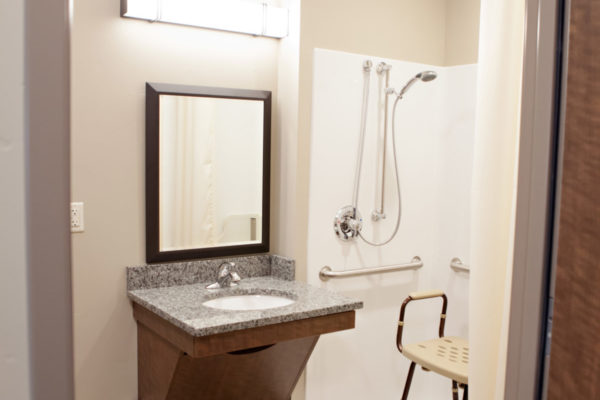 A bathroom with a sink and mirror, a walk-in shower with a stool and grab bars on the wall.