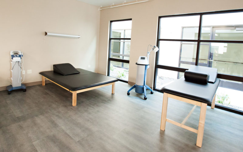 Two therapy tables in a room with large windows. One table is low and wide, the other table is tall and narrow. Both tables have a wedge pillow on them. Two types of therapy equipment on rollers are placed near each bed.