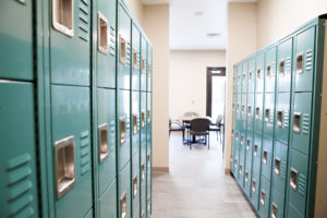 A narrow walkway with four rows of gym-style lockers on each side. The hallway opens into a room with a square table and four chairs.