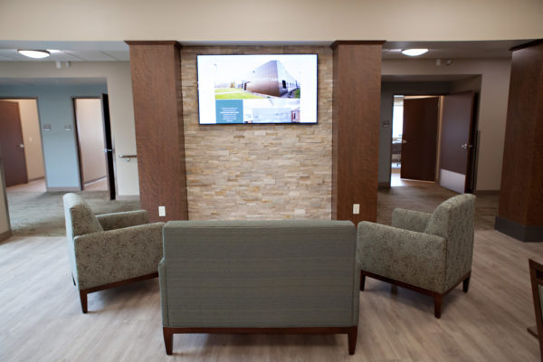 Cascadia of Nampa, Idaho lobby. A love seat and two chairs sit in a semi-circle in front of a brick pillared wall with a wall-mounted television. A large hallway leading to resident rooms is on either side of the wall.